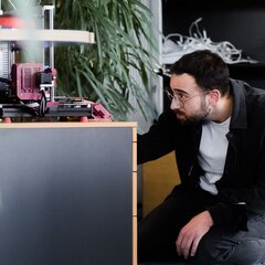 Mann beobachtet einen in Betrieb befindlichen 3D-Drucker in einem kreativen Büroumfeld. | © Photo: Ilja Kagan, 2022
