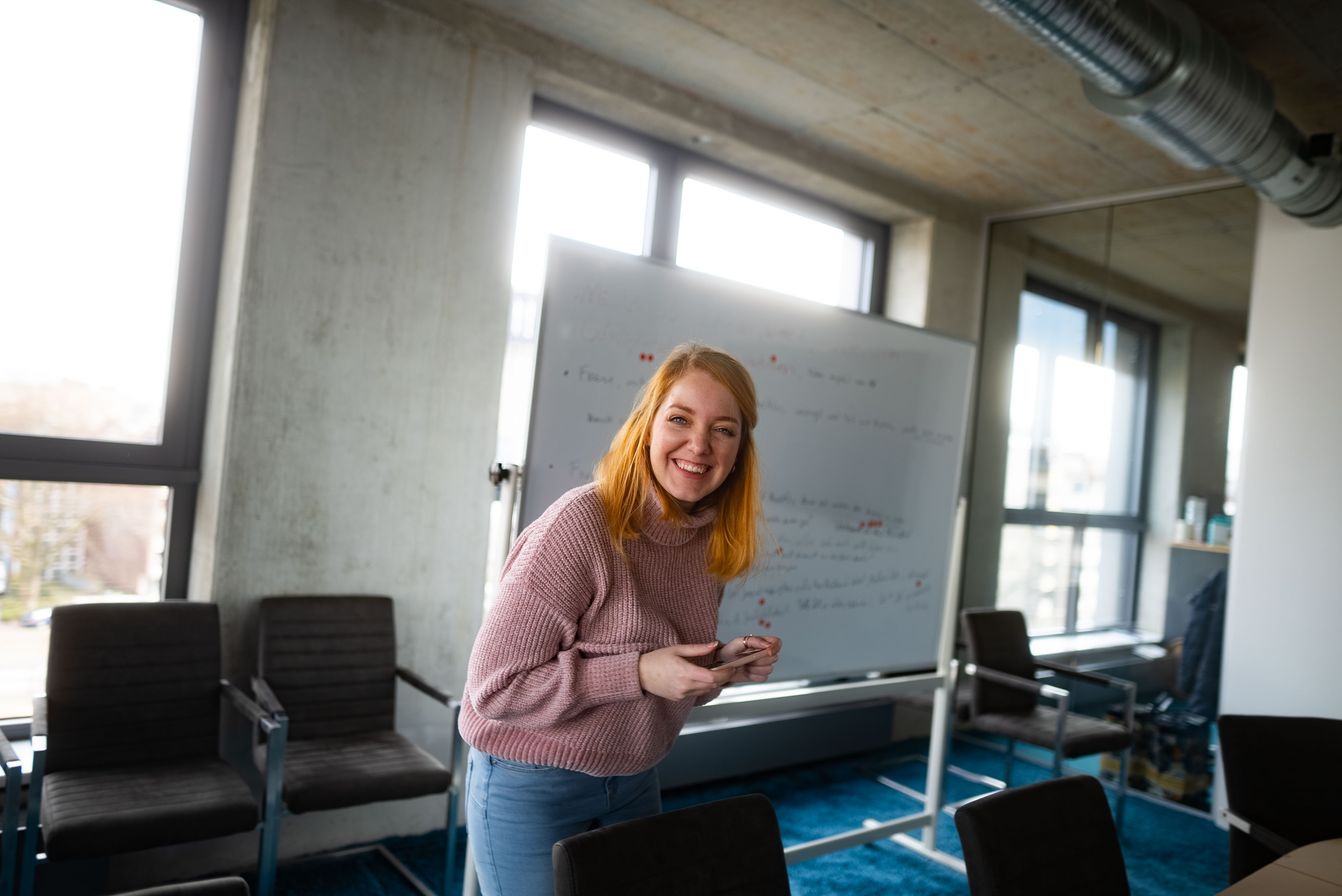 Presentation at whiteboard in office. | © Photo: Ilja Kagan, 2022 A smiling woman stands in front of a whiteboard in a modern office. She is wearing a pink sweater and looking at the camera. | © Photo: Ilja Kagan, 2022