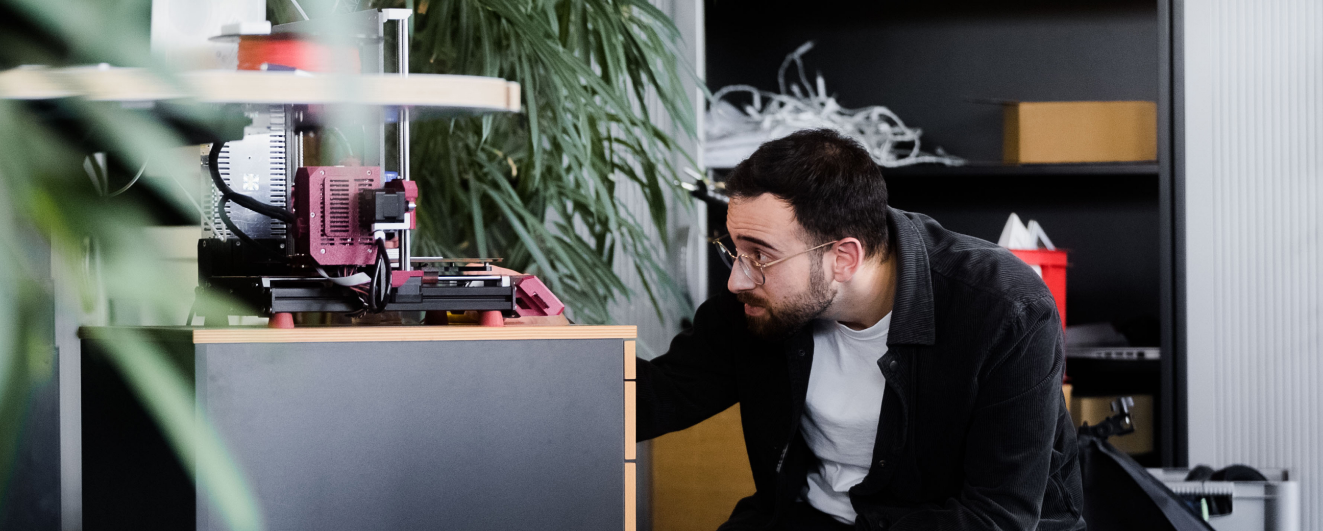 Technician inspecting 3D printer. | © Photo: Ilja Kagan, 2022 Man with glasses kneeling in front of a desk inspecting a 3D printer in the office. | © Photo: Ilja Kagan, 2022