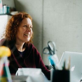 Happy woman with curly red hair smiling while working on her laptop in a modern office environment.